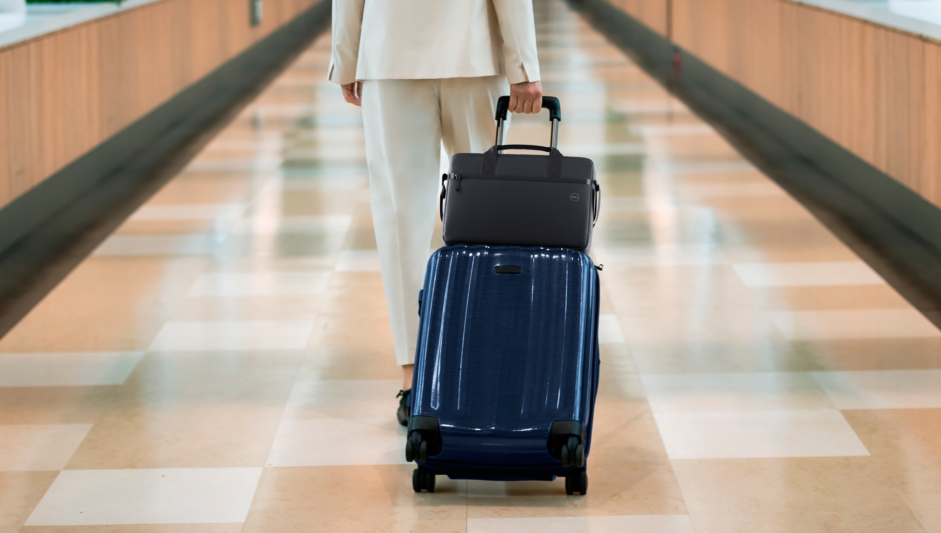 Woman walking down a long hallway carrying a suitcase with a briefcase resting on top of it.