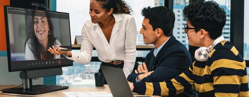 Lenovo ThinkSmart View Plus, with camera and soundbar attached, sitting on a desk in front of three individuals communicating with a young woman via a virtual meeting software