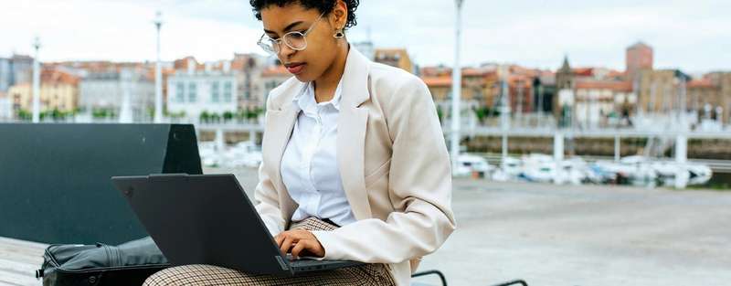 Front view of a professional working on the Lenovo ThinkPad T14s Gen5 (14â Intel) Eclipse Black laptop placed on its lap, sitting outdoors, focusing its carbon-fiber-based top cover & slim profile.