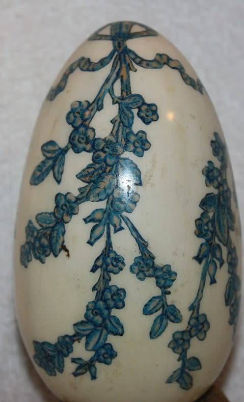 LARGE DECORATIVE CERAMIC EGG . WHITE BACKGROUND WITH BLUE RIBBON AND FLORAL DETAIL ON A LOOSE METAL