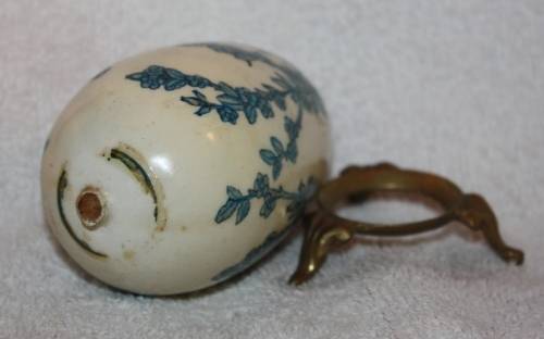 LARGE DECORATIVE CERAMIC EGG . WHITE BACKGROUND WITH BLUE RIBBON AND FLORAL DETAIL ON A LOOSE METAL