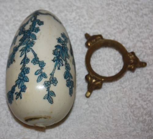 LARGE DECORATIVE CERAMIC EGG . WHITE BACKGROUND WITH BLUE RIBBON AND FLORAL DETAIL ON A LOOSE METAL