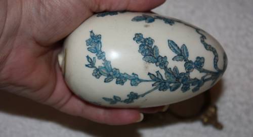 LARGE DECORATIVE CERAMIC EGG . WHITE BACKGROUND WITH BLUE RIBBON AND FLORAL DETAIL ON A LOOSE METAL