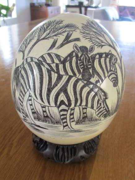 AN OSTRICH EGG ON CARVED WOODEN STAND DEPICTING TWO ZEBRAS DRINKING WATER