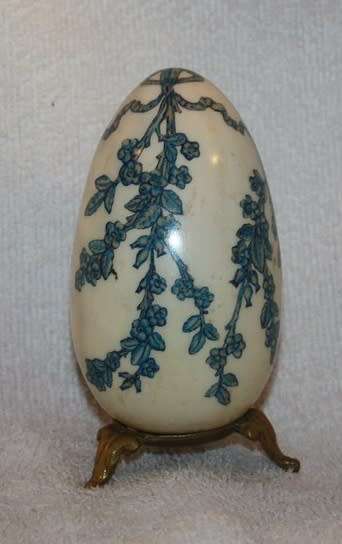 LARGE DECORATIVE CERAMIC EGG . WHITE BACKGROUND WITH BLUE RIBBON AND FLORAL DETAIL ON A LOOSE METAL