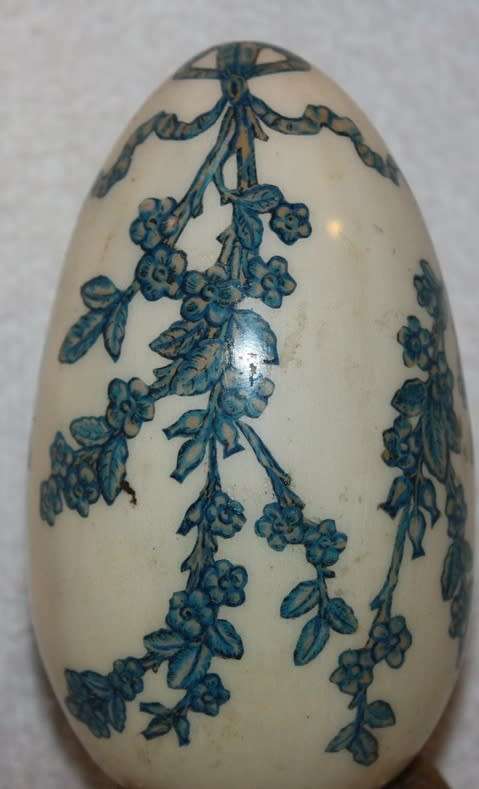 LARGE DECORATIVE CERAMIC EGG . WHITE BACKGROUND WITH BLUE RIBBON AND FLORAL DETAIL ON A LOOSE METAL