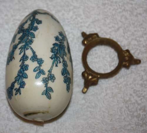 LARGE DECORATIVE CERAMIC EGG . WHITE BACKGROUND WITH BLUE RIBBON AND FLORAL DETAIL ON A LOOSE METAL